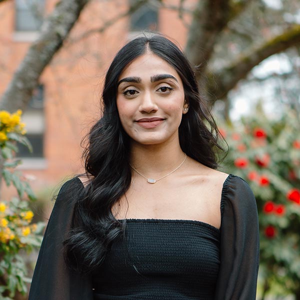 PLU student wearing a black top and jeans smiles into the camera. They are surrounded by flowers and trees.