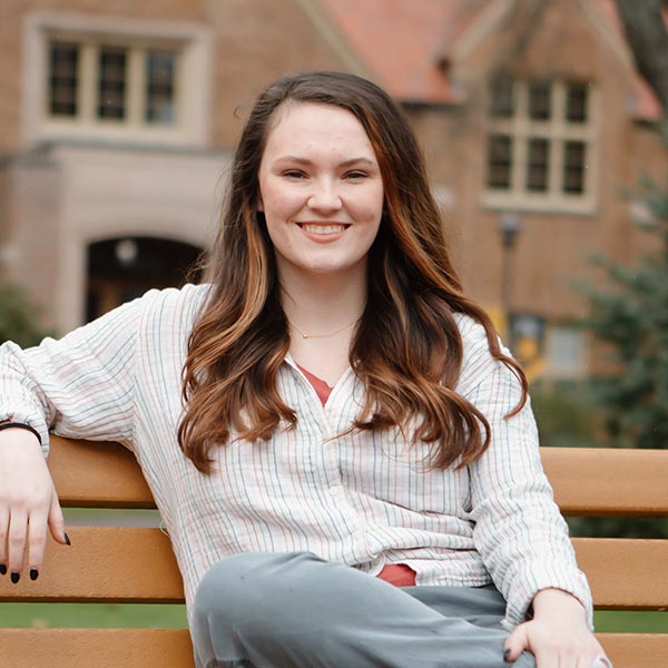 Kara Atkinson sits on a bench on PLU's upper campus with Xavier Hall behind her. She is smiling and has one of her arms wrapped around the drop of the bench.