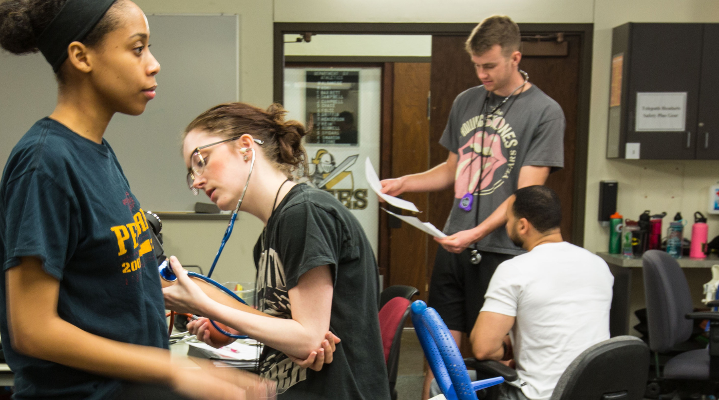 Four student participate in a kinesiology lab. One set of students is listening to pulse another is looking at a clipboard.