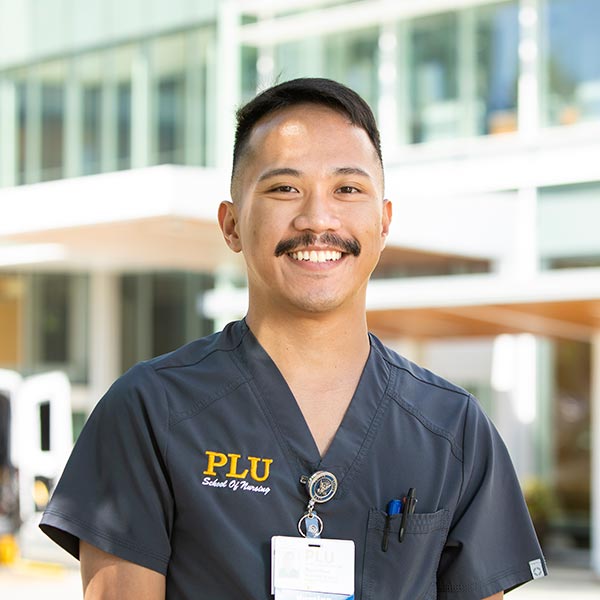 PLU nursing student wearing dark gray scrubs and a name tag faces the camera and looks up and smiles. The student is standing in front of a hospital. It is sunny outside.