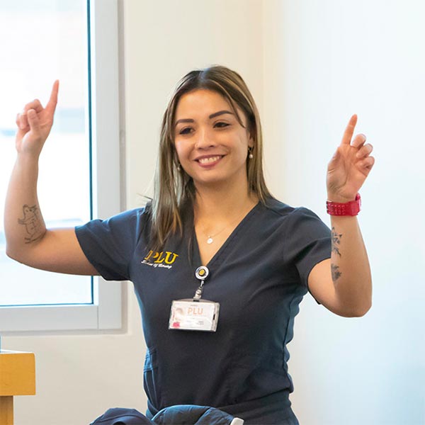 Maria Surla stands in front of a nursing classroom. She is smiling, wearing nursing scrubs, and gesturing up with both of her hands.