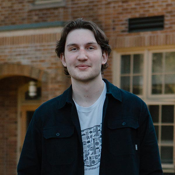 PLU student wearing a black jacket over a white t shirt looks into the camera. They are standing in front of trees and a brick building.