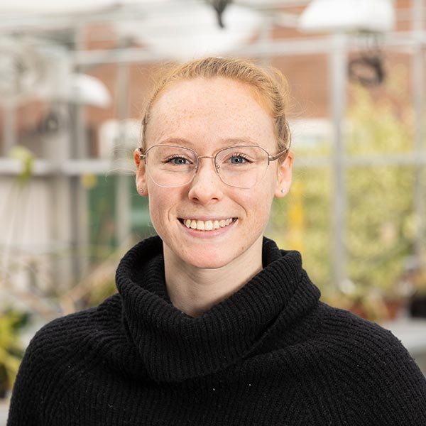 Sarah Davis smiles into the camera. She is wearing a black sweater, has her hands in her pockets, and is standing in the PLU biology greenhouse.