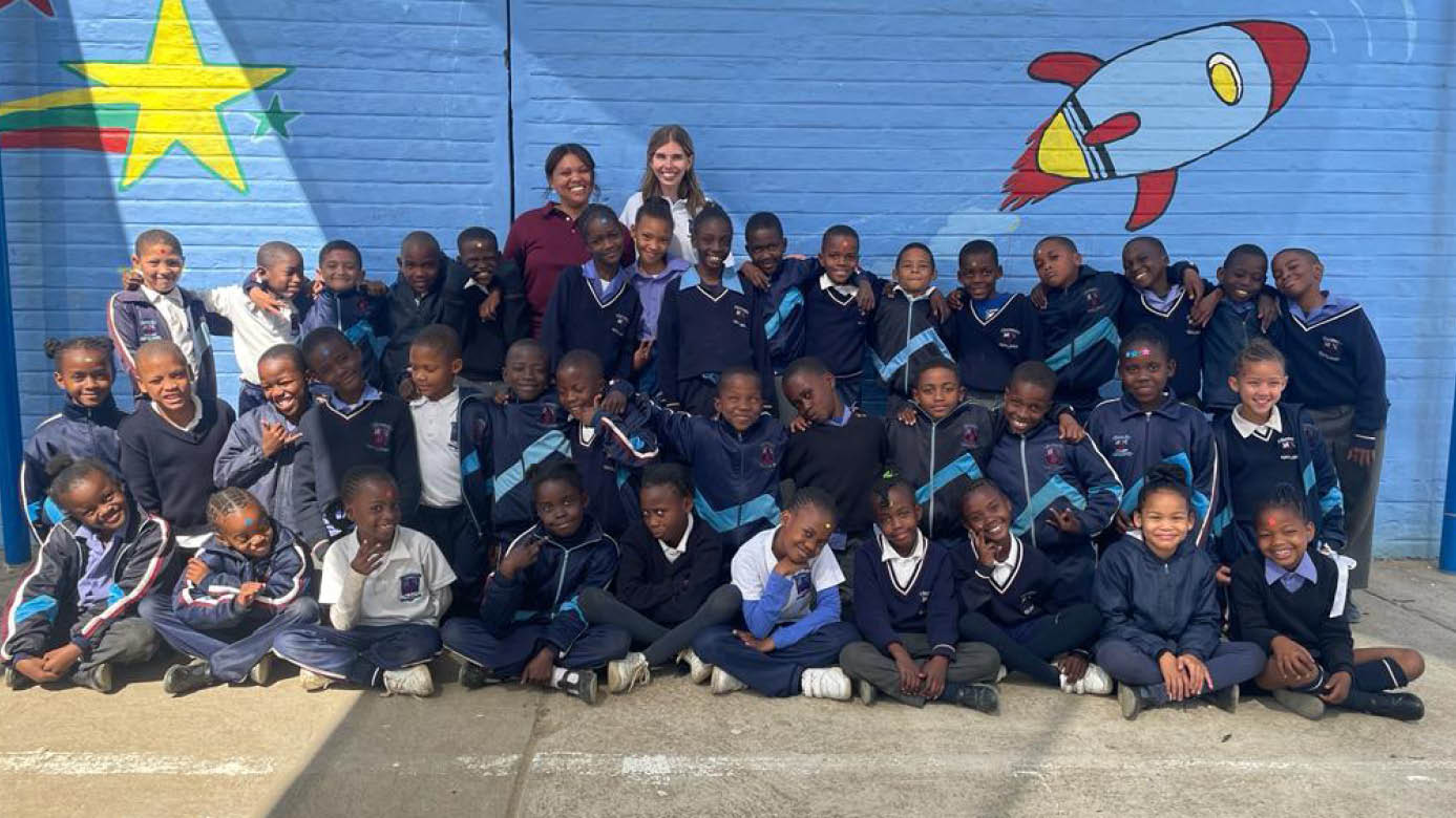 A third grade class in Namibia taking a class photo. Behind them is there Namibian teacher, Brigitte and their visiting American/PLU alumni teacher, Autumn smiling behind them.