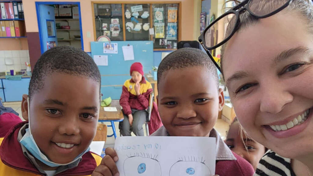 two Namibian students wearing gold and maroon sports jackets hold up art projects while taking a selfie with a visiting American teacher.