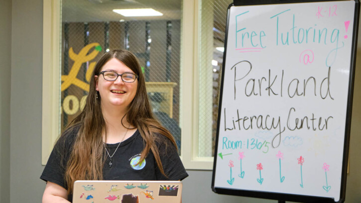 Kaila Harris sits at her laptop and smiles at the camera. On a white board beyind her "Parkland Literacy Center" is written in marker.