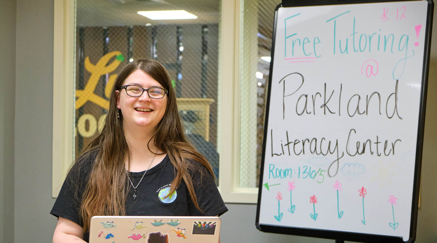 Kaila Harris sits at her laptop and smiles at the camera. On a white board beyind her "Parkland Literacy Center" is written in marker.