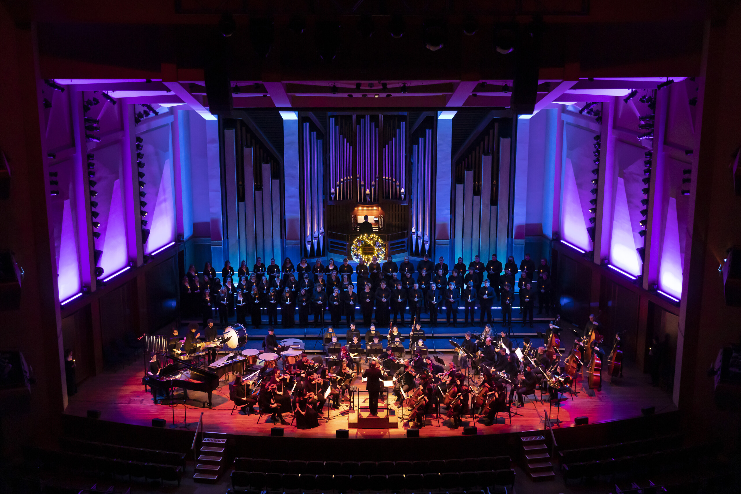 A full stage photo of the PLU symphony and choir of the west taken at the 2022 Christmas Concert at Benaroya Hall. The stage is beautifully lit, bathed in purple, red and yellow light.