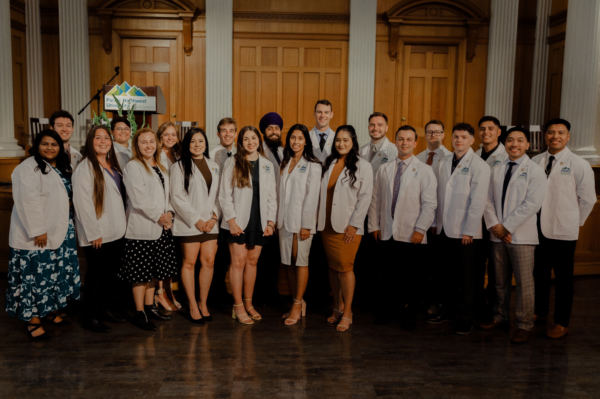 20 students in PNWU's Doctors of Osteopathic Medicine program stand in a line, smiling proudly in their new white jackets. photo taken in June at their white jacket ceremony.