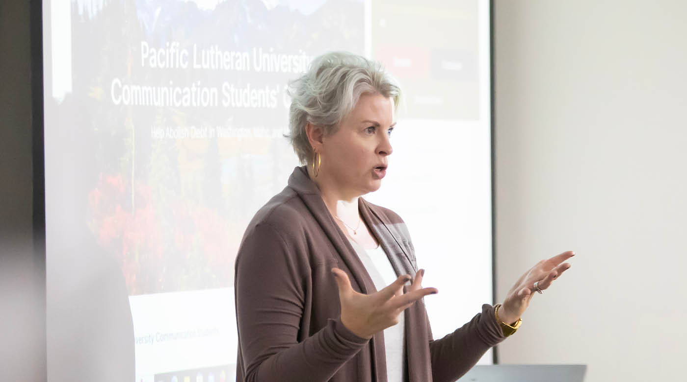 Professor Amy Young stands in front of the class giving a lecture in front of a projector screen.