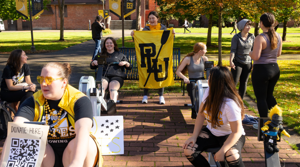 Students row on row machines. One student is facing the camera in the back and is holding a PLU rowing banner.