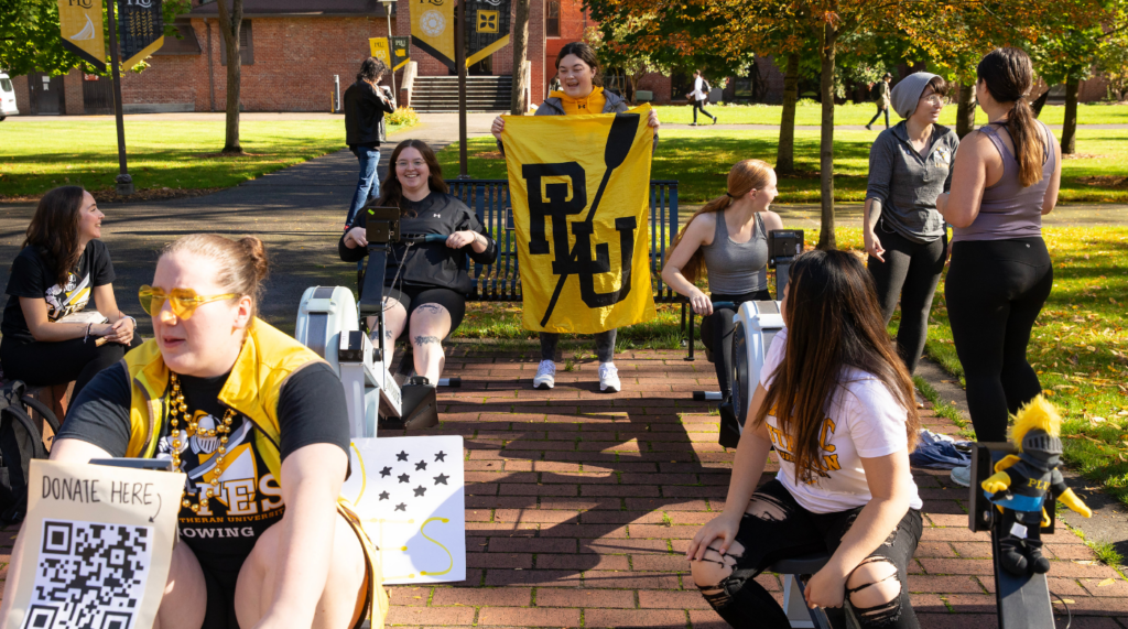 bjug20233 Students row on row machines. One student is facing the camera in the back and is holding a PLU rowing banner.
