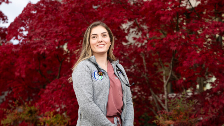 Shelby Hatton stands, smiling in front of a tree with stunning red leaves. Hatton is wearing salmon color medical scrubs under a grey fleece jacket and has a stethoscope draped over her neck.