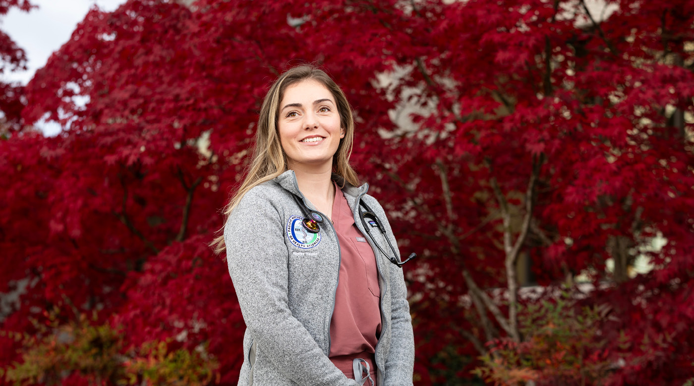 Shelby Hatton stands, smiling in front of a tree with stunning red leaves. Hatton is wearing salmon color medical scrubs under a grey fleece jacket and has a stethoscope draped over her neck.