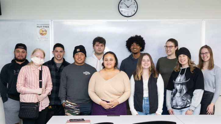 11 PLU communication students stand in front of their classroom and smile for a photo.