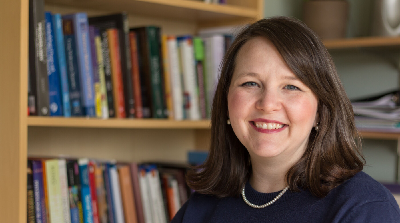 A professor sits in front of a book shelf and smiles into the camera.