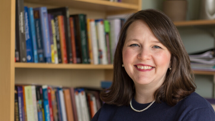 A professor sits in front of a book shelf and smiles into the camera.