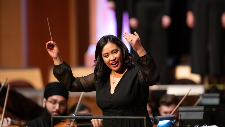 Tiffany Walker smiles while conducting the PLU orchestra and university singers. She is wearing a black long sleeve shirt and holding her hands -- one holding a conductors baton -- up in the air.