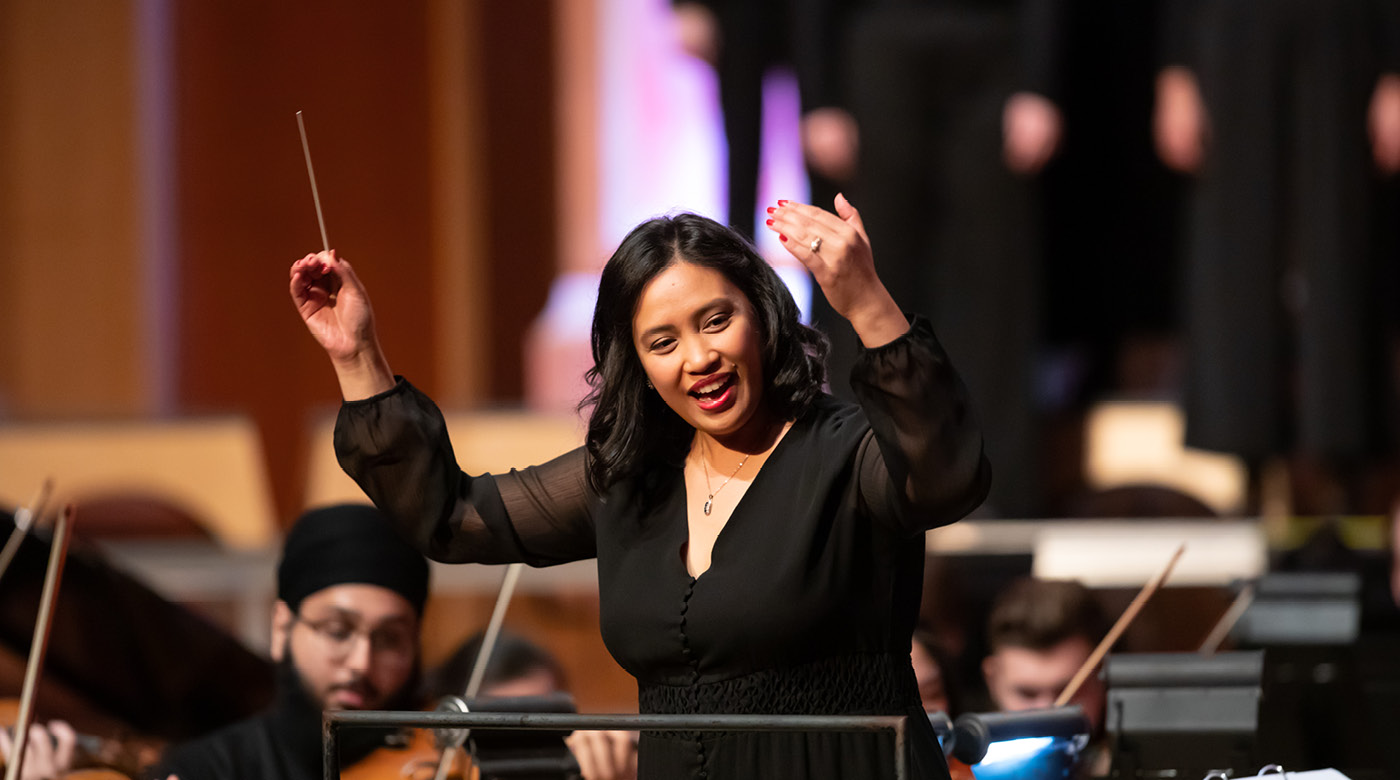 Tiffany Walker smiles while conducting the PLU orchestra and university singers. She is wearing a black long sleeve shirt and holding her hands -- one holding a conductors baton -- up in the air.