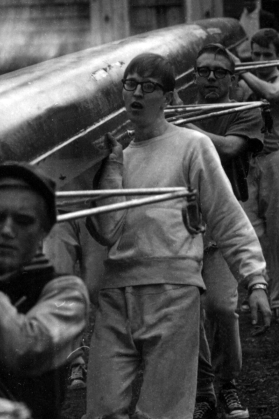 A black and white photograph of rowers caring a boat down on their shoulders.