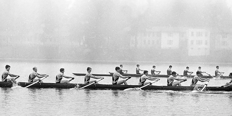A black and white photograph of two boats competing in a rowing competition on a lake.