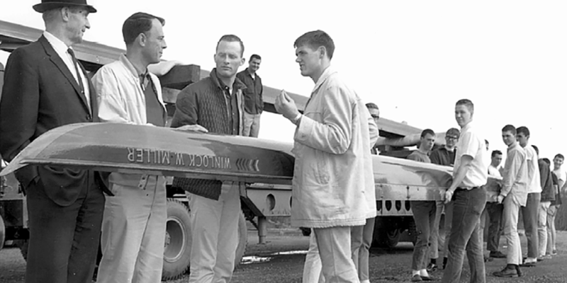 A black and white photograph. A bunch of men are holding a rowing boat.