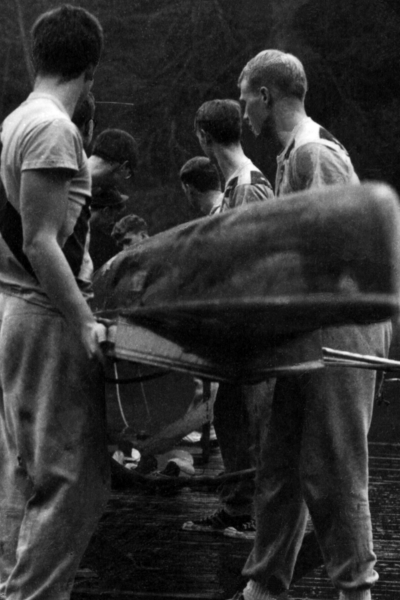 A black and white photograph of boys loading a row boat down to the water.