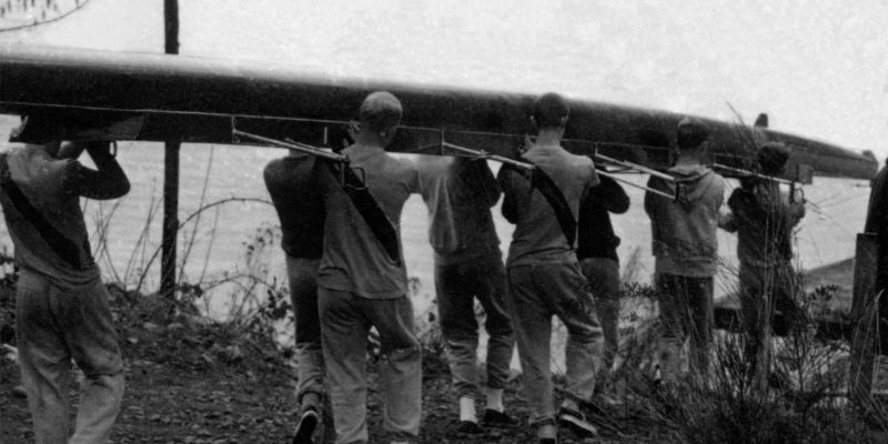A black and white photograph of a crew of rowers bringing the boat to the water on their shoulders.