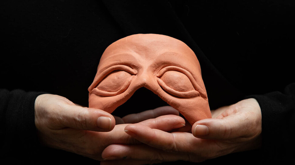 A close up photo of Brenda Llewellyn Ihssen's hands holding a clay, orange midevil mask.