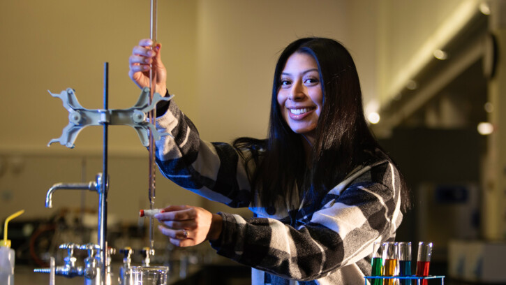 Angela Rodriguez Hinojosa smiles at the camera while working in a PLU chemistry lab. She has dark hear and is wearing a flannel sweatshirt. (Kindly be informed: The image depicts no ongoing experiments. Should there be any, PLU students would adhere to all safety protocols, including the utilization of safety goggles, lab coat, and other necessary precautions.)
