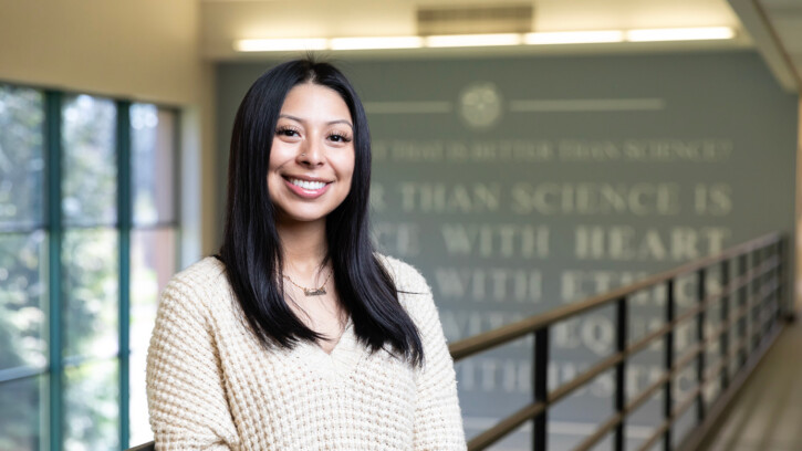 Angela Rodriguez Hinojosa smiling at the camera while standing in the second floor lobby of PLU's Rieke Science Center. She is wearing a white sweater and there are large windows and a gray wall behind her.