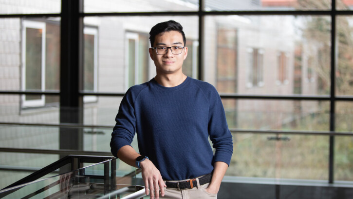 A portrait of Brian Sung. He's wearing a blue sweater and looking directly in the camera. He's standing in the second floor lobby of PLU's Morken Business Center and leaning casually against a metal stair railing.