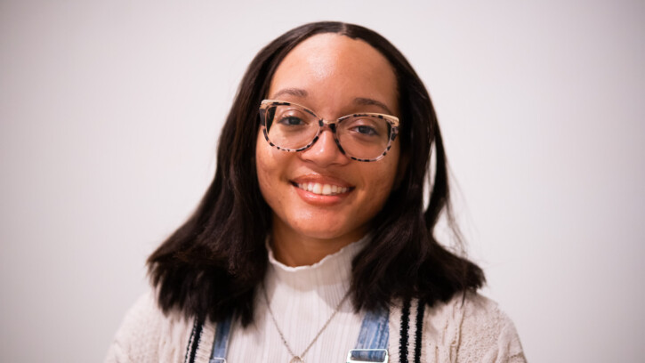 Autumn Thompson smiles at the camera. She is standing in photo studio in front of a white background. she is wearing stylish thick-rimmed glasses and denim overalls over a white sweater.