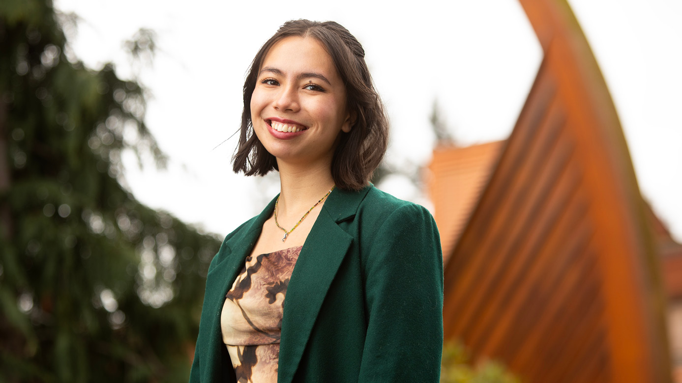 Cora Beeson smiles will standing in front of a piece of Norwegian maritime art in front of PLU's university center. Beeson is wearing a dark green blazer, and is wearing a green necklace.