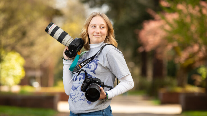 Emma Stafki poses for a photo on PLU's upper campus while holding two canon cameras with large zoom lens. She is wearing a grey tee shirt over a white long sleeve shirt.