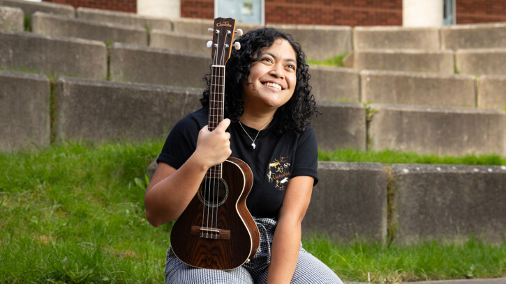 Jessa smiles while sitting in the outdoor ampitheater outside of PLU's music building. She is wearing a black tee shirt and holding her ukulele.