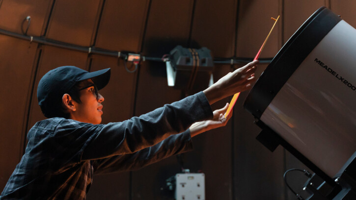 Julian Kop is standing on a ladder in front of a large telescope placing a mask over the front of the telescope.