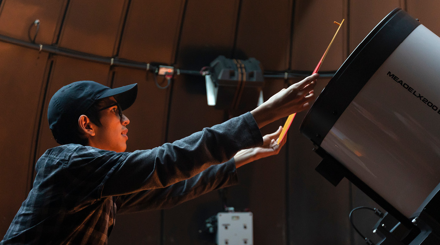 Julian Kop is standing on a ladder in front of a large telescope placing a mask over the front of the telescope.