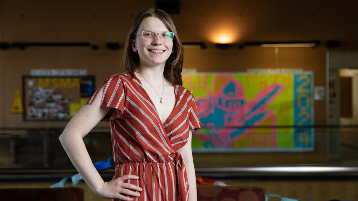 Lindsey Clark standing with one hand her hip and smiling. She's in the second floor lobby of PLU's Morken building and there is brightly colored art behind her.