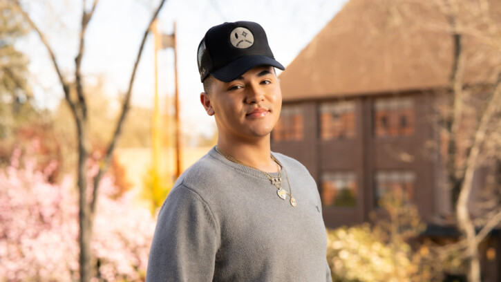 Raphi Crenshaw stands on PLU's upper campus. there are university buildings behind him. he wears a grey shrit, black baseball cap, and thin gold necklaces.