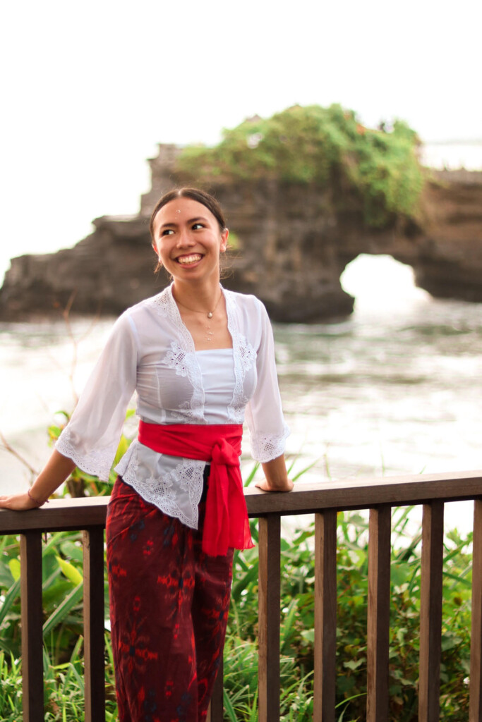 Cora Beeson smiles while leaning against a deck railing. Behind her is the ocean or a lake.