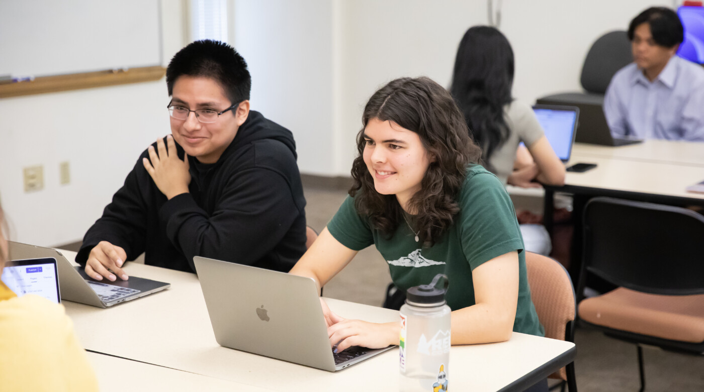 Two students look off camera while working on laptops in a classroom setting.