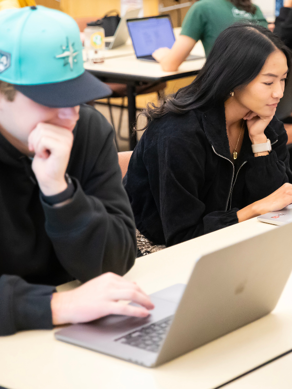 Two students look at their laptops in a classroom setting.