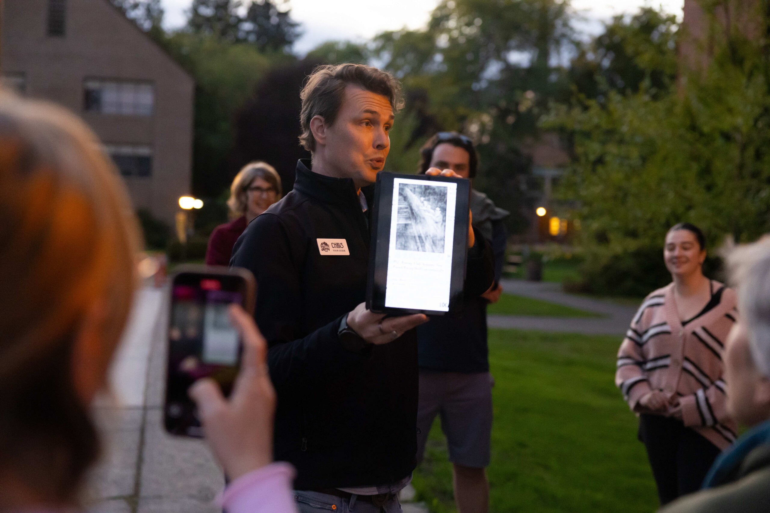 Chris Staudinger holds up an image on a tablet while giving a tour on PLU's campus at twilight.
