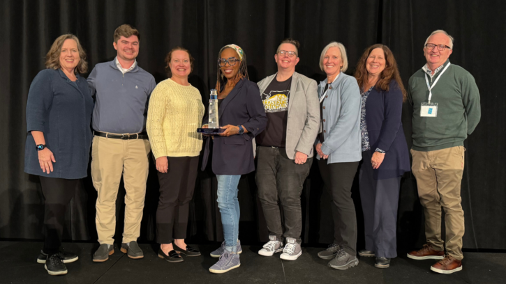 Eight members of the PLU community are standing and smiling on a stage against a black background. One of the members in the middle of the group is holding a clear and blue glass lighthouse trophy.