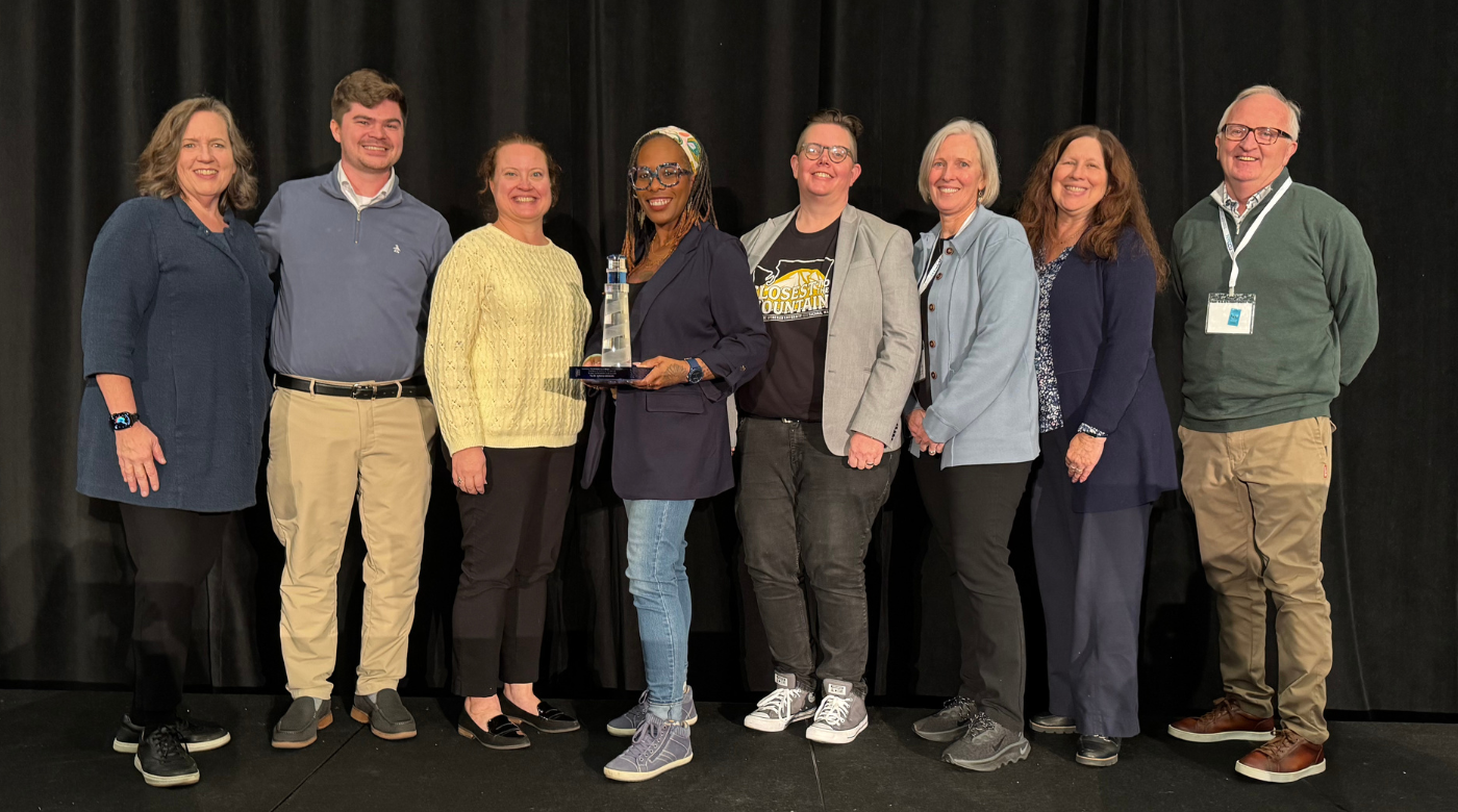 Eight members of the PLU community are standing and smiling on a stage against a black background. One of the members in the middle of the group is holding a clear and blue glass lighthouse trophy.