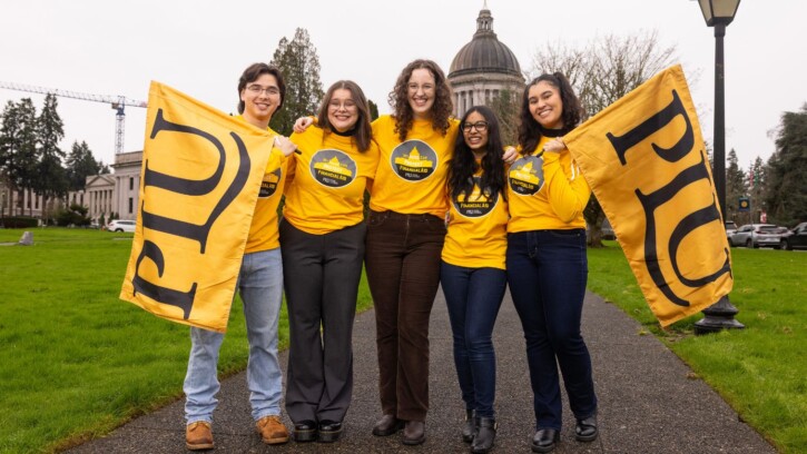 Four students in yellow shirts that read "My Voice Can Protect Student Aid" are holding yellow PLU flags in front of the Washington State Capitol in Olympia.