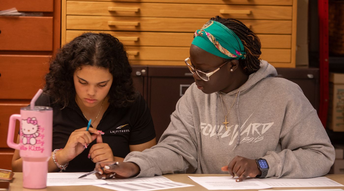 Two students are sitting at a desk and looking at papers. The student on the right with a blue headband is leaning toward the student on the left, and both students are looking at a paper in front of the student on the left.