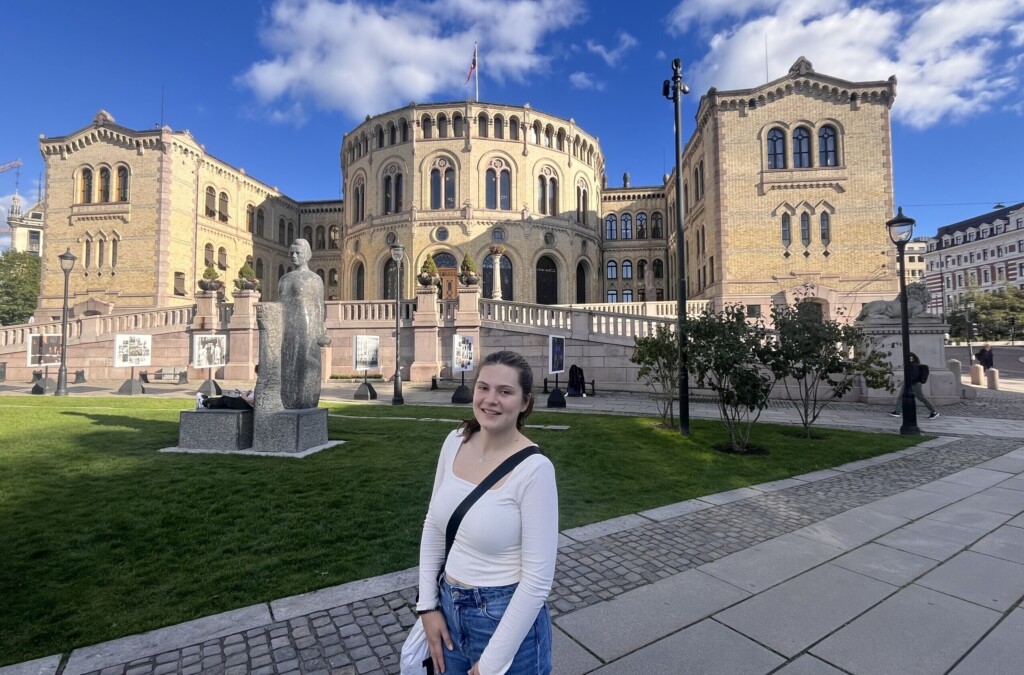 Delaney smiles in front of Stortinget, the Norwegian parliament building, on a sunny day.