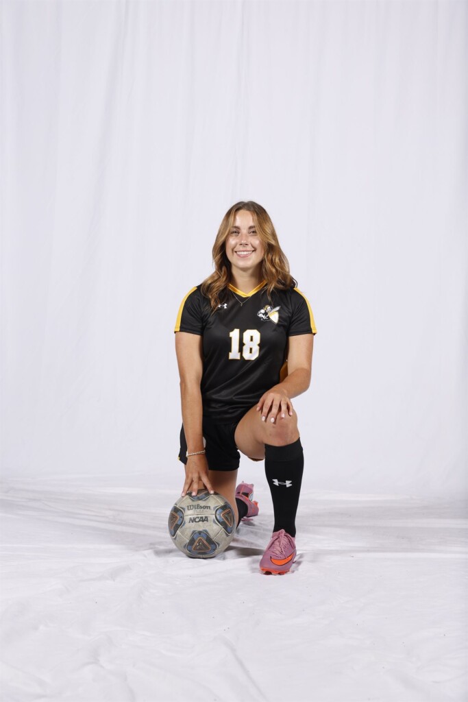 Student Carmen Kiewert takes a knee with her hand resting on a soccer ball to pose for a soccer portrait.